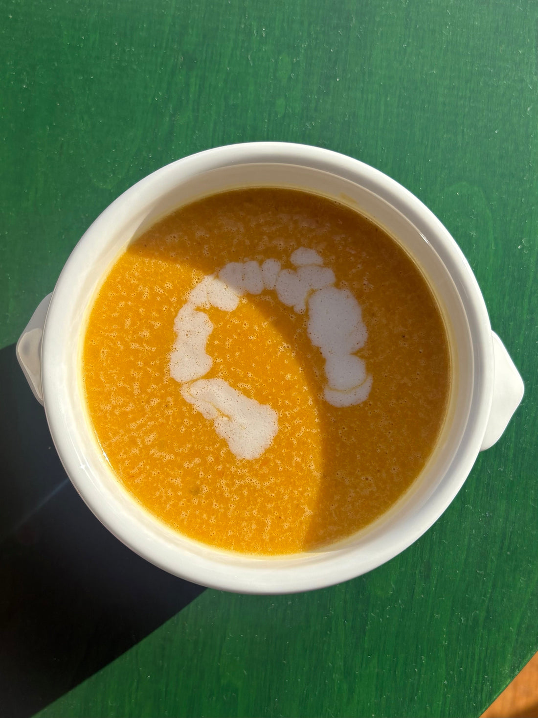 Butternut squash soup in a white bowl, garnished with a drizzle of coconut milk, served on a green table.