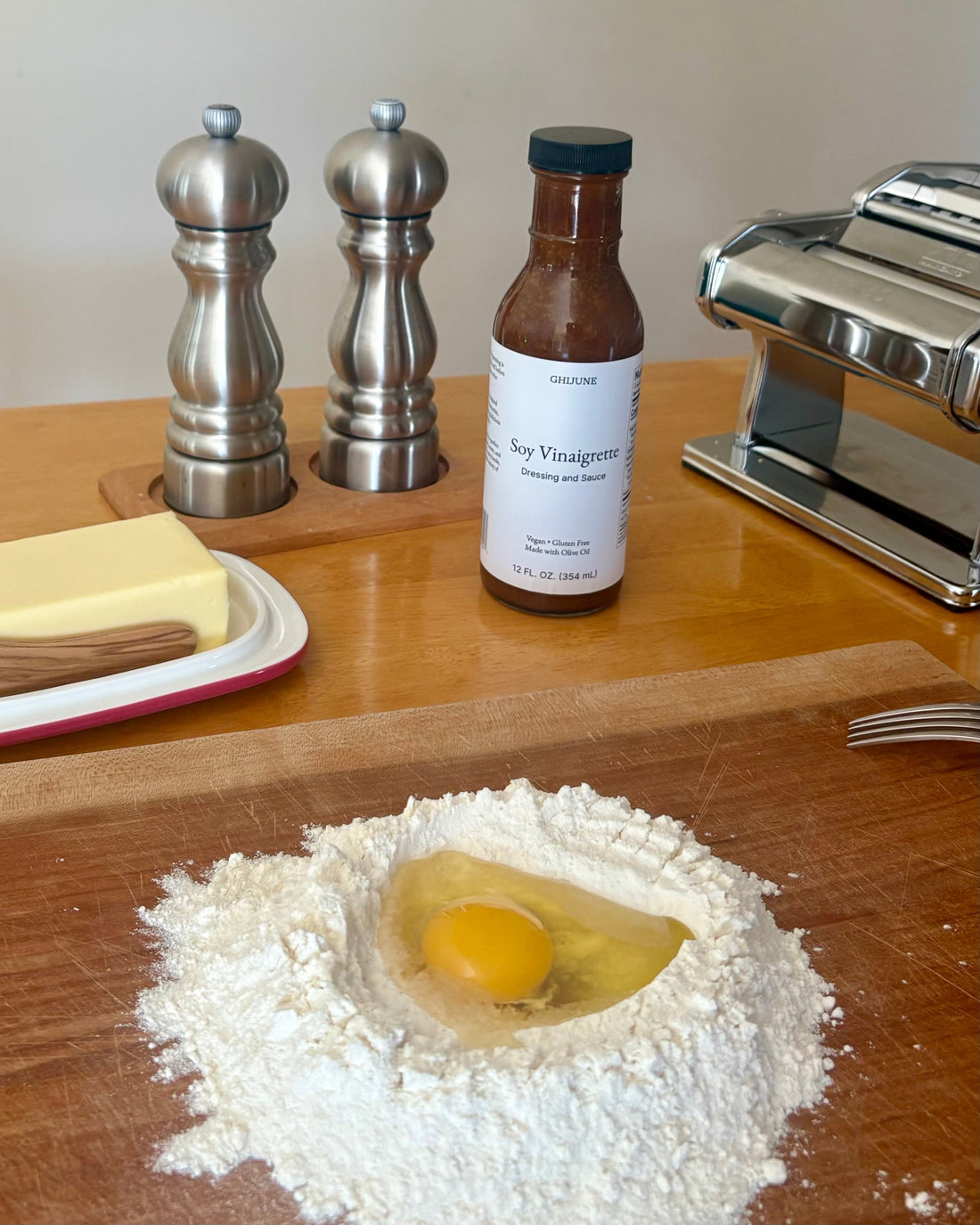 Ingredients and tools for making fresh homemade pasta arranged on a countertop, including flour with an egg in the center, butter, a butter knife, salt and pepper shakers, GHIJUNE Soy Vinaigrette Salad Dressing & Sauce, a pasta maker, and a fork.