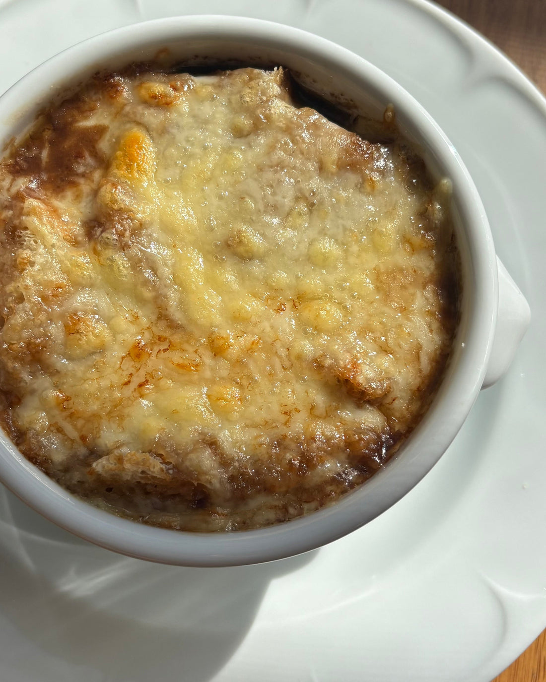 A bowl of caramelized onion soup topped with toasted bread and melted golden cheese, served in an oven-safe bowl with a rich, savory broth.