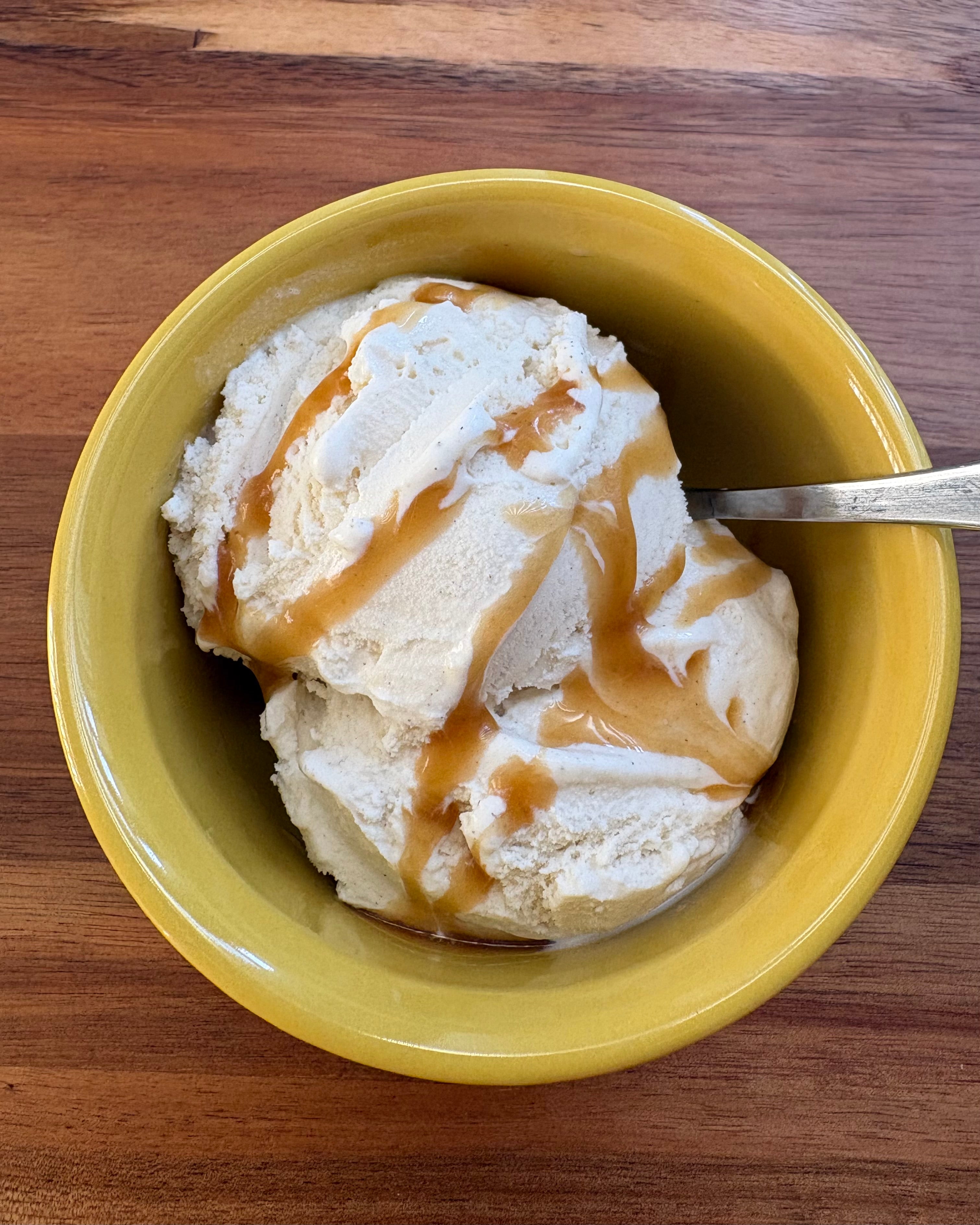 A yellow bowl filled with scoops of vanilla ice cream drizzled with a glossy honey soy sauce. A spoon is inserted into the ice cream, ready to serve, highlighting the creamy texture and rich caramel-colored drizzle.
