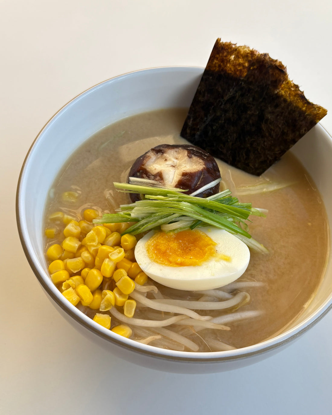 A bowl of GHIJUNE miso noodle soup topped with seaweed, corn, bean sprouts, a half-boiled egg, green onions, and shiitake mushrooms, with noodles and savory broth visible beneath the colorful toppings.