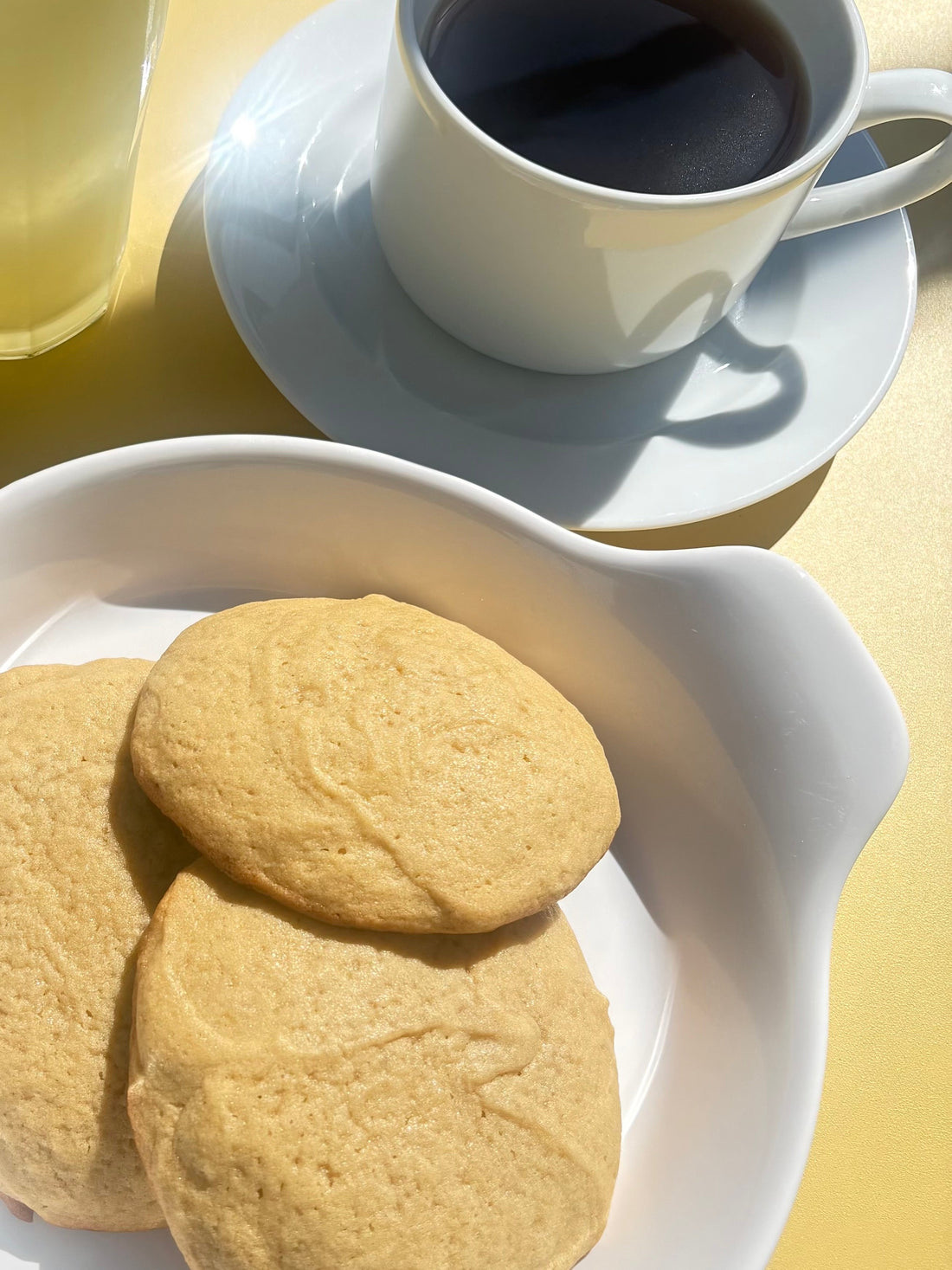 A yellow tabletop holds a glass of lemonade, a cup of coffee on a matching saucer, and a white plate with three butter cookies. The simple arrangement creates a bright, inviting café-style scene.