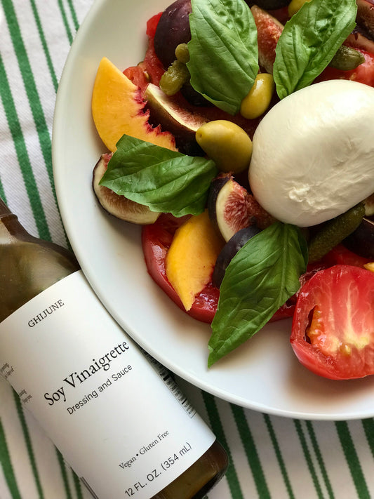 a plate of heirloom tomatoes, figs, basil, burrata cheese, peach, olive, and cornichons, and a condiment bottle. 